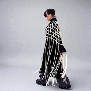 May include: A black coat with a white macrame overlay and fringe detail. The coat is worn with dark gray pants and black boots. The macrame design creates a geometric pattern across the shoulders and back.
