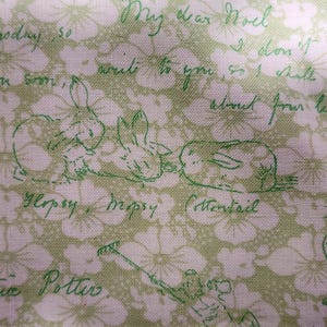 May include: A light green fabric featuring a repeating white floral pattern overlaid with dark green line drawings of rabbits and cursive script text that includes the names Flopsy, Mopsy, Cottontail, and the signature of Beatrix Potter.