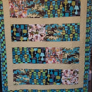 May include: A quilt with a patchwork design featuring a variety of fabrics. The quilt is made up of squares and rectangles of different colors and patterns, including camouflage, guitars, and abstract designs. The quilt is bordered with a blue and green fabric with a guitar pattern.