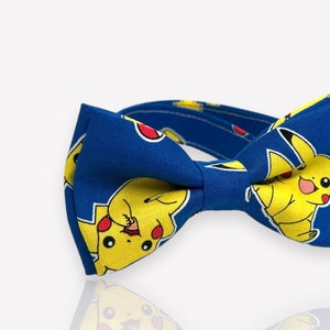 May include: A blue bow tie featuring a repeating pattern of a yellow cartoon character with red cheeks and a lightning bolt tail. The bow tie is made of fabric and has a pre-tied bow shape.