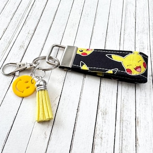 Könnte beinhalten: Ein schwarzer und gelber Stoffschlüsselanhänger mit einem lächelnden Pikachu-Charakter. Der Schlüsselanhänger hat einen silbernen Metallring und einen gelben Smiley-Anhänger mit einem gelben Quasten.