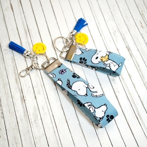 Könnte beinhalten: Zwei hellblaue Schlüsselanhänger mit Snoopy-Muster. Jeder Schlüsselanhänger hat einen silbernen Metallverschluss, einen gelben Smiley-Anhänger und eine blaue Quaste. Das Snoopy-Muster zeigt Snoopy, Pfotenabdrücke und Blumen.