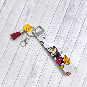 Puede incluir: Un llavero de tela blanca con un diseño de Mickey Mouse rojo, amarillo y negro. El llavero tiene un anillo de metal plateado y un borla roja.