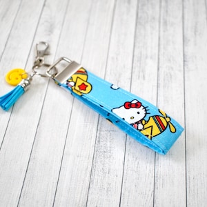 May include: A blue fabric keychain wristlet featuring a Hello Kitty design with a red bow and yellow accents. The keychain includes a silver-tone metal clasp, a yellow smiley face charm, and a blue tassel.