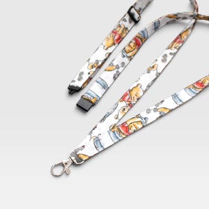 May include: A white fabric lanyard featuring a repeating watercolor-style print of a yellow bear wearing a red shirt, interspersed with small blue and grey floral accents. The lanyard includes a black plastic adjustable slider and a silver metal clasp.