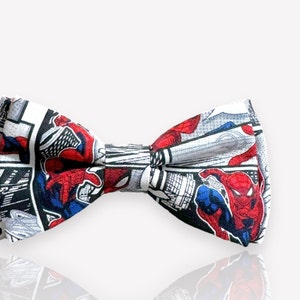 Peut inclure: Un nœud papillon blanc avec un imprimé de bande dessinée représentant Spiderman en rouge et bleu. Le nœud papillon a un fond blanc avec des contours noirs.