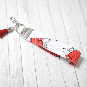 Puede incluir: Un llavero de muñeca con temática de Snoopy en blanco y rojo con una borla roja. La tela presenta a Snoopy en su caseta. El llavero tiene un cierre de metal plateado y una correa para la muñeca.