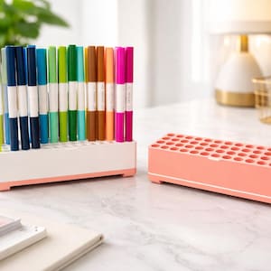 Könnte beinhalten: Eine Sammlung farbenfroher Dual-Tip-Marker in einem weiß-rosa Organizer. Daneben steht ein separater, passender rosa Markerhalter. Die Marker sind nach Farben sortiert, darunter Gelb, Blau, Grün und Pink.