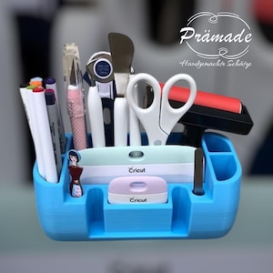 Könnte beinhalten: Ein blauer Bastel-Organizer mit verschiedenen Werkzeugen, darunter Stifte, Scheren und ein Cricut-Rollschneider. Der Organizer hat mehrere Fächer und enthält ein Cricut-Etikettiergerät. Der Text "Prämade Handgemachte Schätze" ist sichtbar.
