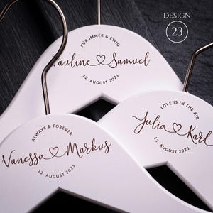 Puede incluir: Tres perchas de madera blanca con ganchos de metal en oro rosa. Cada percha presenta texto personalizado en marrón oscuro y un gráfico de corazón. El texto incluye nombres como Pauline & Samuel, Julia & Karl y Vanessa & Markus, con fechas y frases.