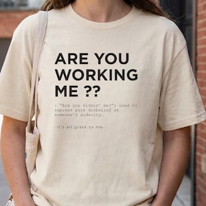 Puede incluir: Camiseta color crema con el texto "ARE YOU WORKING ME ??" en negro. La camiseta incluye una definición de la frase y la frase "it's all greek to me." La persona lleva un gorro marrón y vaqueros azules.