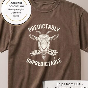 Può includere: T-shirt marrone con grafica di capra e le parole "Predictably Unpredictable". La maglietta è tinta in capo e realizzata in materiale pesante. La grafica presenta una testa di capra con la parola "Caution" su uno striscione.