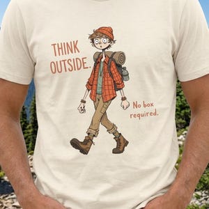 May include: Cream-colored t-shirt with a graphic of a cartoon hiker. The hiker wears a red plaid shirt, brown boots, and a backpack. Text on the shirt reads "THINK OUTSIDE. No box required."