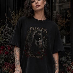May include: A black short-sleeve t-shirt featuring a graphic print of a cloaked figure and ethereal spirits. The text "THANATOS" appears at the top, with "THE LAST WITNESS" printed below the central illustration in a muted color palette.
