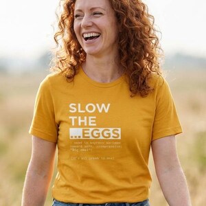 Może przedstawiać: Musztardowa koszulka z białym napisem "SLOW THE ...EGGS". Koszulka jest połączona z niebieskimi dżinsami. Osoba idzie po polu wysokiej trawy.