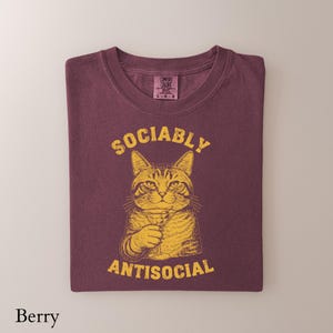 May include: Berry-colored t-shirt with a graphic of a cat holding a cocktail glass. The words "Sociably" and "Antisocial" are printed above and below the cat illustration in a yellow font. The shirt is folded.