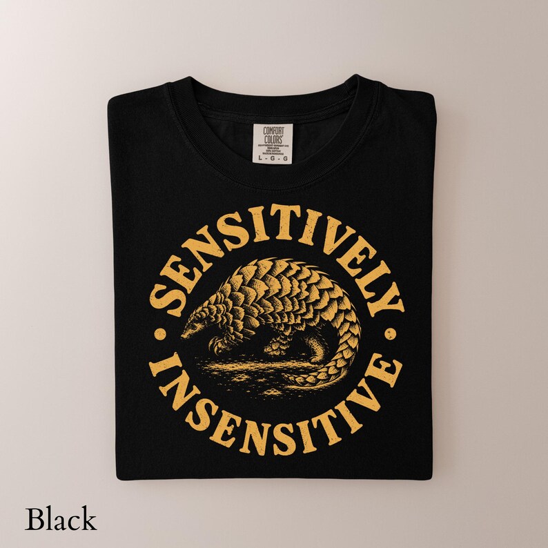 May include: A black t-shirt featuring a yellow graphic of a pangolin centered within a circular border. The text "SENSITIVELY INSENSITIVE" is printed in a bold, arched font around the animal. The shirt has a Comfort Colors brand neck label.