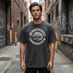 May include: A charcoal gray short-sleeve t-shirt featuring a circular graphic on the chest. The design includes a detailed illustration of a pangolin in the center, surrounded by the text SENSITIVELY INSENSITIVE in a bold, white, sans-serif font.