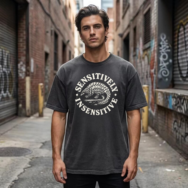 May include: A charcoal gray short-sleeve t-shirt featuring a circular graphic on the chest. The design includes a detailed illustration of a pangolin in the center, surrounded by the text SENSITIVELY INSENSITIVE in a bold, white, sans-serif font.