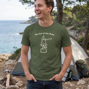 May include: Olive green t-shirt with the text "The Art of the Brew" and a white graphic of a coffee pot on a portable stove. The shirt is being worn outdoors near a tent and the ocean.