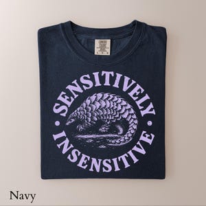 May include: A navy blue t-shirt featuring a light purple graphic of a pangolin centered within a circular frame. The text "SENSITIVELY INSENSITIVE" is printed in a distressed, arched font around the pangolin. The shirt has a Comfort Colors label.