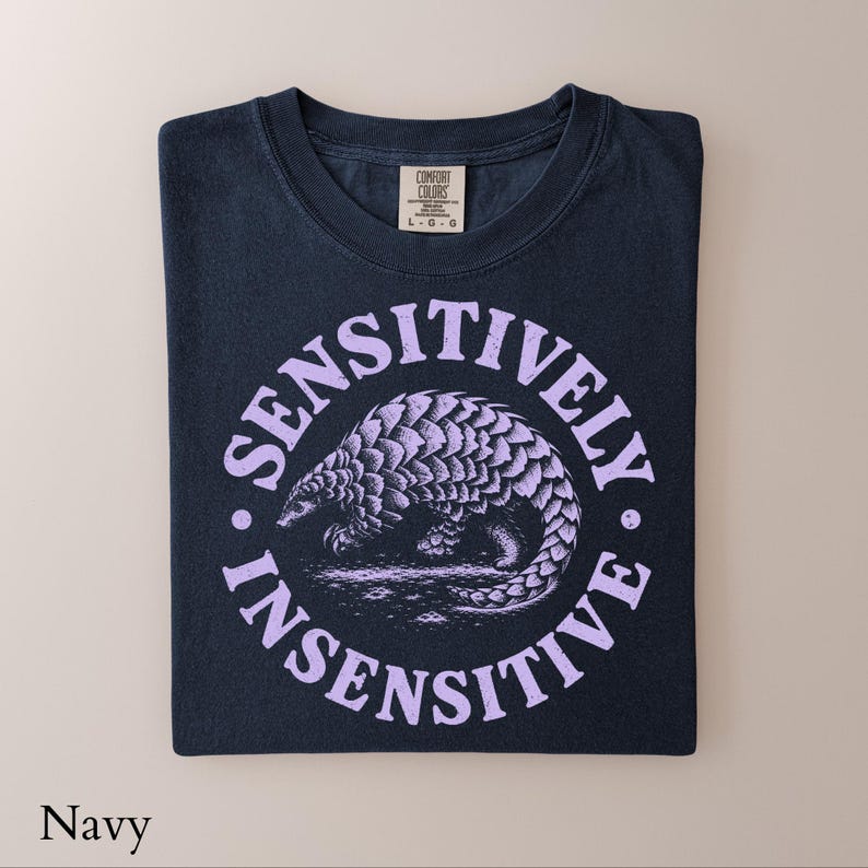May include: A navy blue t-shirt featuring a light purple graphic of a pangolin centered within a circular frame. The text "SENSITIVELY INSENSITIVE" is printed in a distressed, arched font around the pangolin. The shirt has a Comfort Colors label.