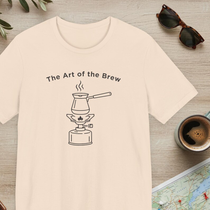 May include: Cream-colored t-shirt with the text "The Art of the Brew" above a black line drawing of a coffee pot on a burner. Sunglasses and a cup of coffee are visible on a wooden surface.