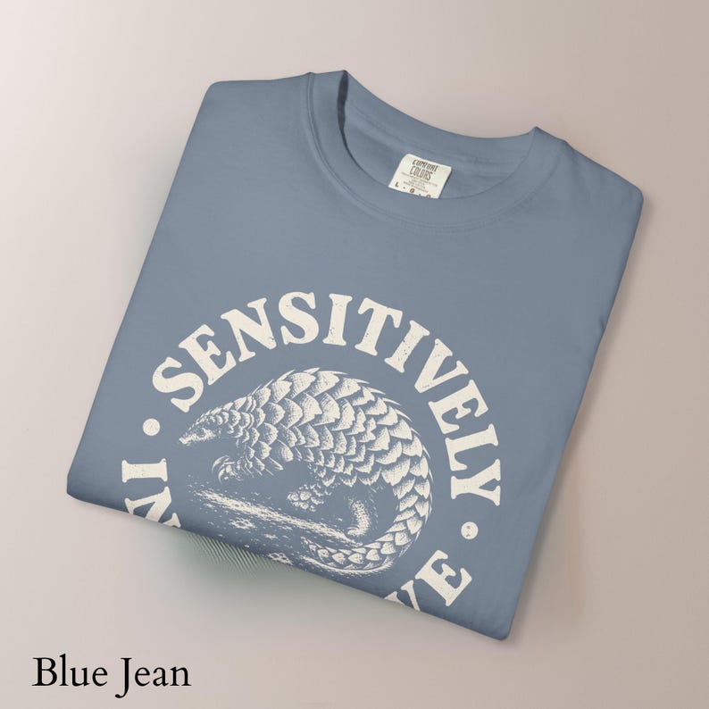 May include: A blue jean colored t-shirt featuring a white graphic of a curled pangolin centered within a circular frame. The text "LIVE SENSITIVELY" is printed in a bold, arched font around the pangolin illustration. The shirt has a crew neckline.