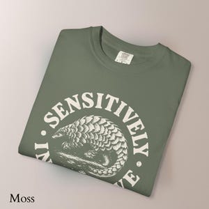 May include: A moss green t-shirt featuring a white circular graphic of a pangolin in the center. The text "LIVE SENSITIVELY" is printed in a distressed white font around the circular border. The shirt has a crew neckline and a Comfort Colors label.