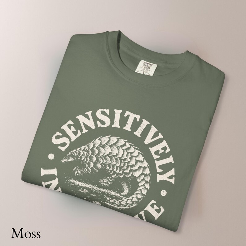 May include: A moss green t-shirt featuring a white circular graphic of a pangolin in the center. The text "LIVE SENSITIVELY" is printed in a distressed white font around the circular border. The shirt has a crew neckline and a Comfort Colors label.