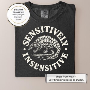 May include: A charcoal gray t-shirt featuring a white circular graphic of a pangolin in the center. The text "SENSITIVELY INSENSITIVE" is printed in a circular arc around the pangolin. The shirt has a Comfort Colors 1717 label at the neck.