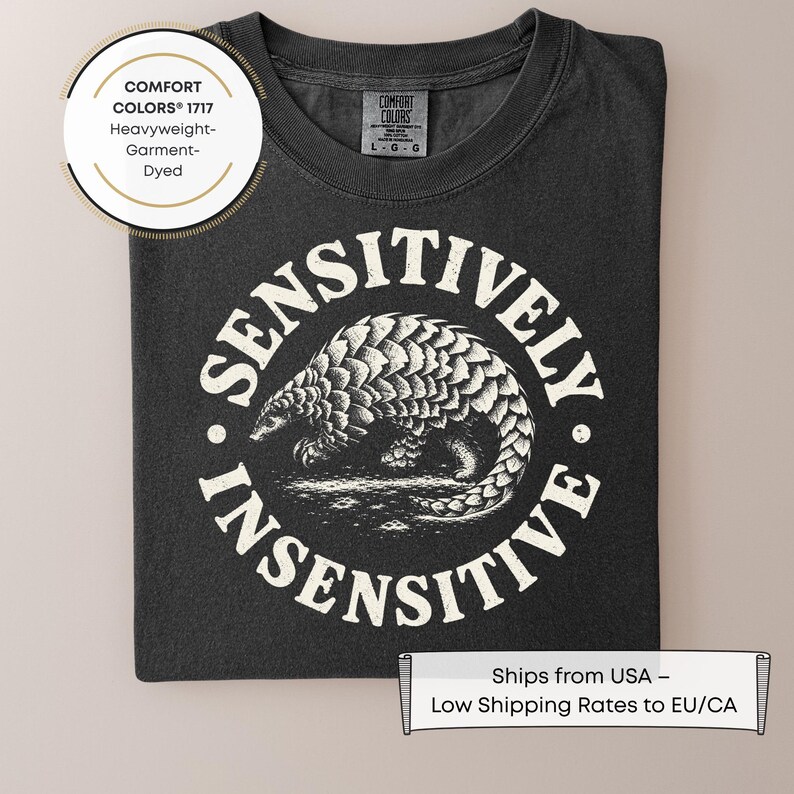 May include: A charcoal gray t-shirt featuring a white circular graphic of a pangolin in the center. The text "SENSITIVELY INSENSITIVE" is printed in a circular arc around the pangolin. The shirt has a Comfort Colors 1717 label at the neck.