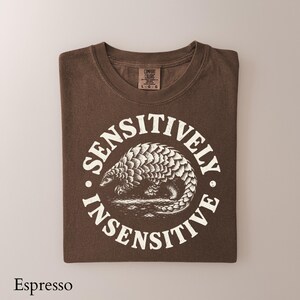 May include: A dark brown short-sleeve t-shirt featuring a white circular graphic of a pangolin in the center. The text "SENSITIVELY INSENSITIVE" is printed in a circular arc around the animal. The shirt has a Comfort Colors brand label.