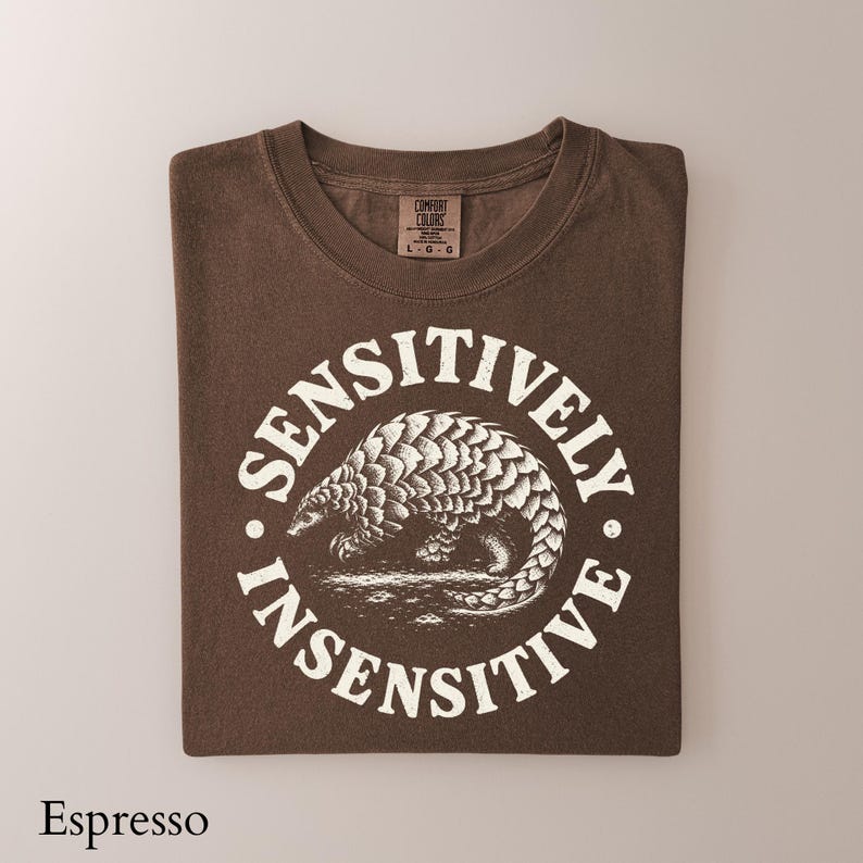 May include: A dark brown short-sleeve t-shirt featuring a white circular graphic of a pangolin in the center. The text "SENSITIVELY INSENSITIVE" is printed in a circular arc around the animal. The shirt has a Comfort Colors brand label.