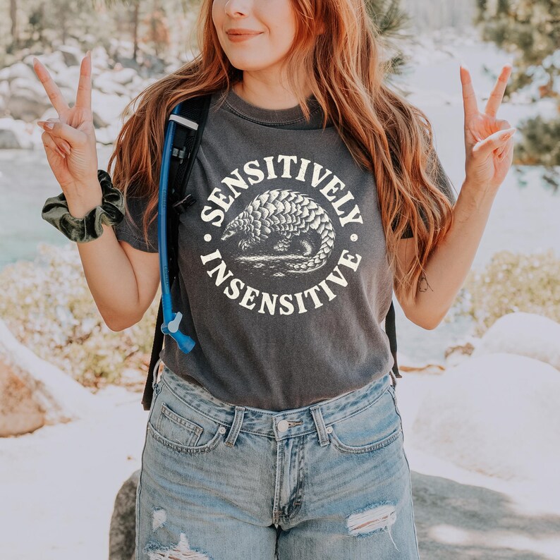 May include: A charcoal gray short-sleeve t-shirt featuring a circular graphic with a pangolin illustration in the center. The text "SENSITIVELY INSENSITIVE" is printed in a white arched font around the pangolin graphic on the front of the shirt.