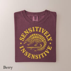 May include: A berry-colored t-shirt featuring a yellow circular graphic with a pangolin illustration in the center. The text SENSITIVELY INSENSITIVE is printed in a circular pattern around the pangolin. The shirt has a Comfort Colors brand label.
