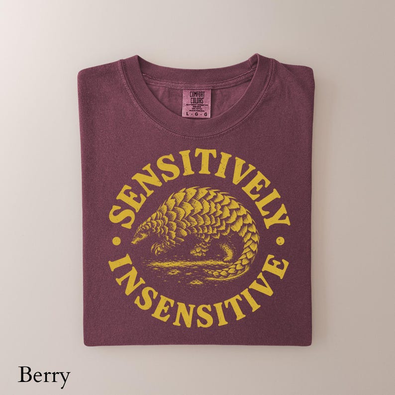 May include: A berry-colored t-shirt featuring a yellow circular graphic with a pangolin illustration in the center. The text SENSITIVELY INSENSITIVE is printed in a circular pattern around the pangolin. The shirt has a Comfort Colors brand label.