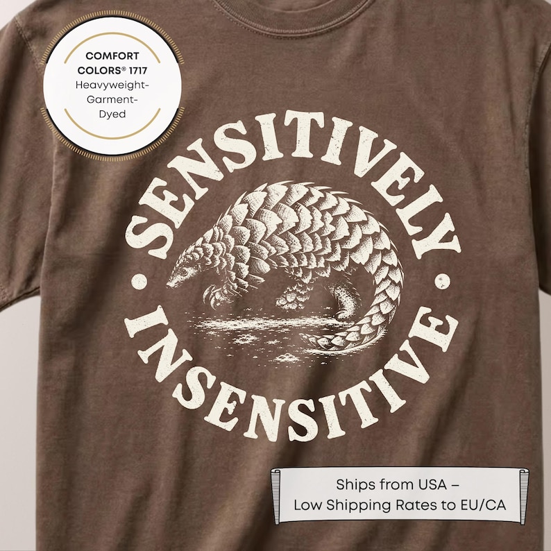 A close-up photograph of a brown Comfort Colors 1717 heavyweight garment-dyed t-shirt. The shirt features a circular white graphic with a detailed, vintage engraving-style illustration of a pangolin covered in sharp protective scales. Distressed block text curves around the animal, reading "SENSITIVELY" along the top half and "INSENSITIVE" along the bottom half.