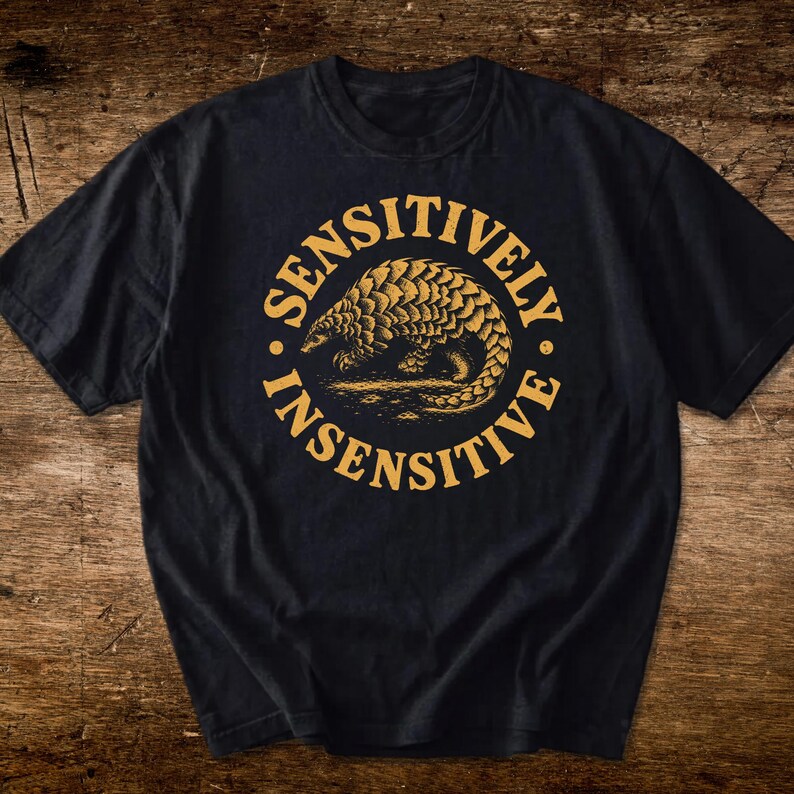 May include: A black short-sleeved t-shirt featuring a circular graphic in mustard yellow. The design depicts a pangolin in the center, surrounded by the text SENSITIVELY INSENSITIVE arranged in a circular border around the animal illustration.