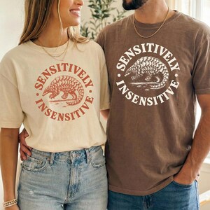 May include: Two short-sleeve crew neck t-shirts. The left shirt is cream with a rust-colored graphic of a pangolin inside a circle with the text SENSITIVELY INSENSITIVE. The right shirt is brown with a white graphic of a pangolin and the same text.