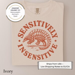 May include: An ivory t-shirt featuring a circular graphic in burnt orange. The design depicts a pangolin in the center, surrounded by the text "SENSITIVELY INSENSITIVE" in a bold, distressed font. The shirt has a Comfort Colors 1717 label.