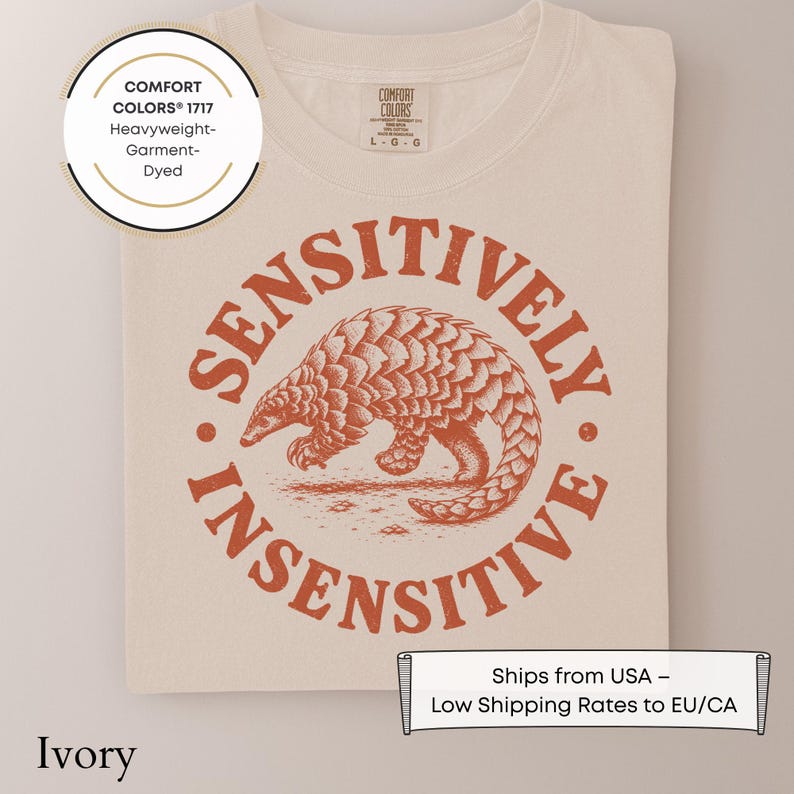 May include: An ivory t-shirt featuring a circular graphic in burnt orange. The design depicts a pangolin in the center, surrounded by the text "SENSITIVELY INSENSITIVE" in a bold, distressed font. The shirt has a Comfort Colors 1717 label.