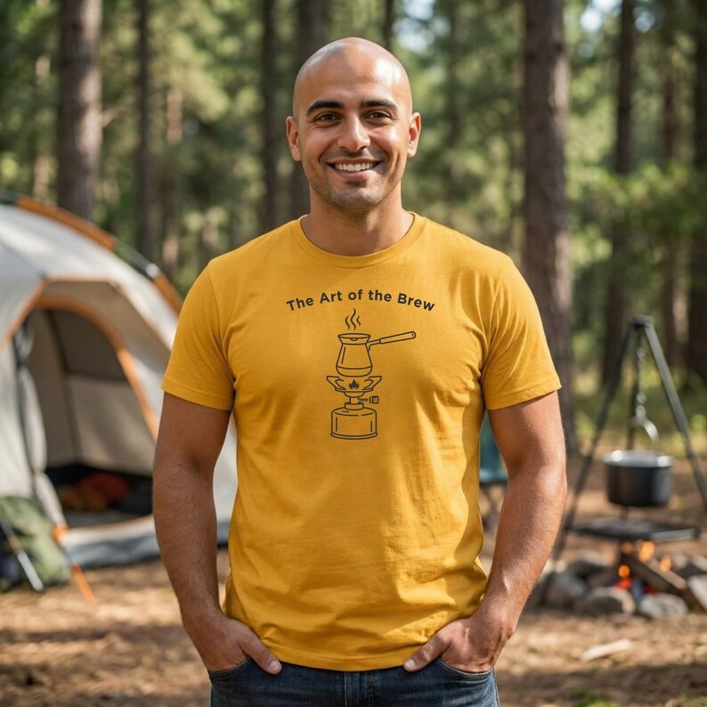May include: Mustard yellow t-shirt with the text "The Art of the Brew" and a black graphic of a coffee pot over a portable stove. The image is taken outdoors with a tent and trees in the background.