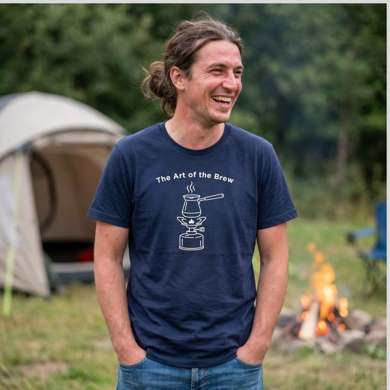 May include: Navy blue t-shirt with the text "The Art of the Brew" and a white graphic of a coffee pot over a portable stove. The person is smiling and standing outside near a tent and a campfire.