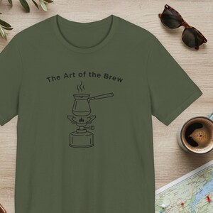 May include: Olive green t-shirt with the text "The Art of the Brew" above a line drawing of a coffee pot on a portable burner. Sunglasses and a cup of coffee are visible in the background.