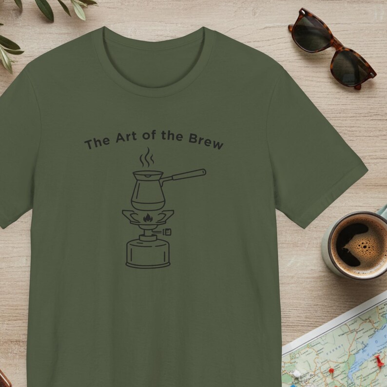 May include: Olive green t-shirt with the text "The Art of the Brew" above a line drawing of a coffee pot on a portable burner. Sunglasses and a cup of coffee are visible in the background.