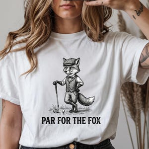 May include: White t-shirt featuring a cartoon fox dressed in golf attire, holding a golf club. The fox is standing on grass with a golf ball. The text "PAR FOR THE FOX" is printed below the fox.
