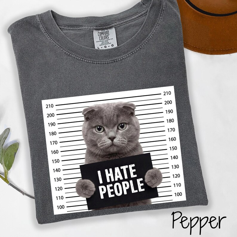 May include: A gray Comfort Colors t-shirt featuring a Scottish Fold cat holding a sign that reads "I HATE PEOPLE." The shirt has a crew neck and short sleeves. The cat is positioned in front of a height chart.