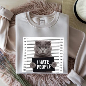 May include: A light beige sweatshirt featuring a gray cat holding a sign that reads "I HATE PEOPLE." The cat is positioned in front of a police lineup background. The sweatshirt has a crew neck and long sleeves.