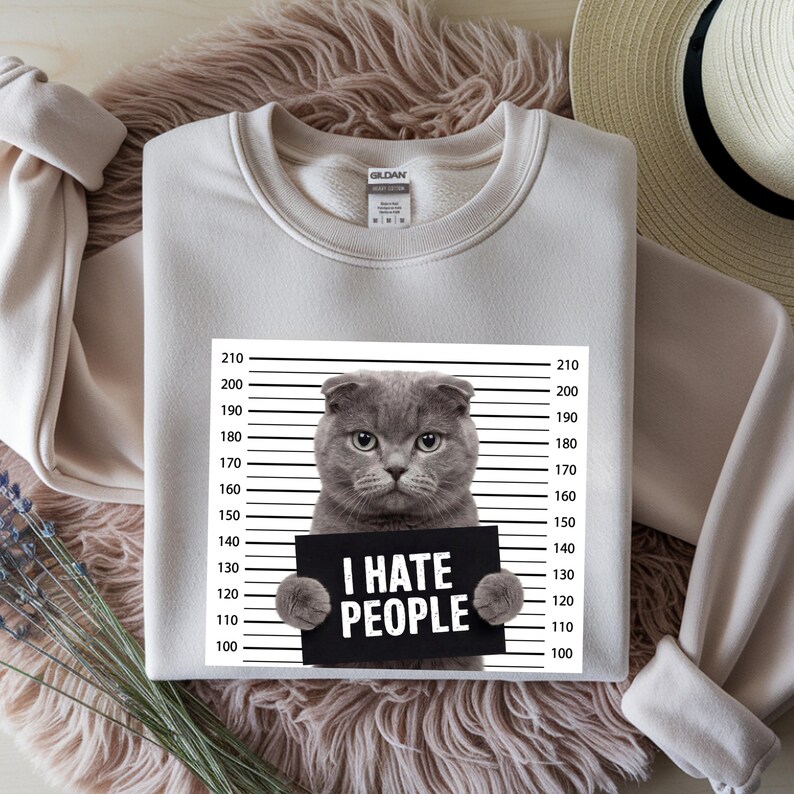 May include: A light beige sweatshirt featuring a gray cat holding a sign that reads "I HATE PEOPLE." The cat is positioned in front of a police lineup background. The sweatshirt has a crew neck and long sleeves.